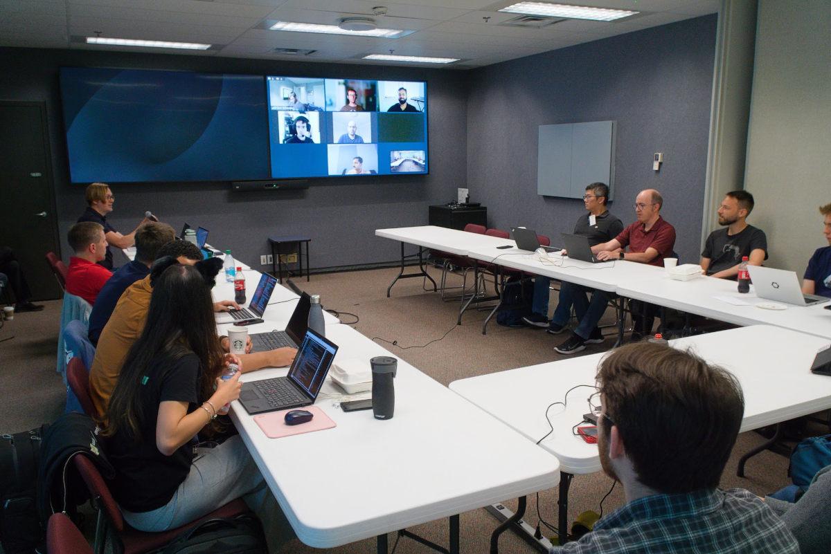 A bunch of people with laptops sitting at desks facing each other. A screens in the background with remote attendees.